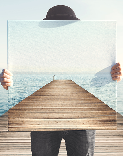 a man stands on a long pier, holding in front of him an image of the pier behind him, as an illusion